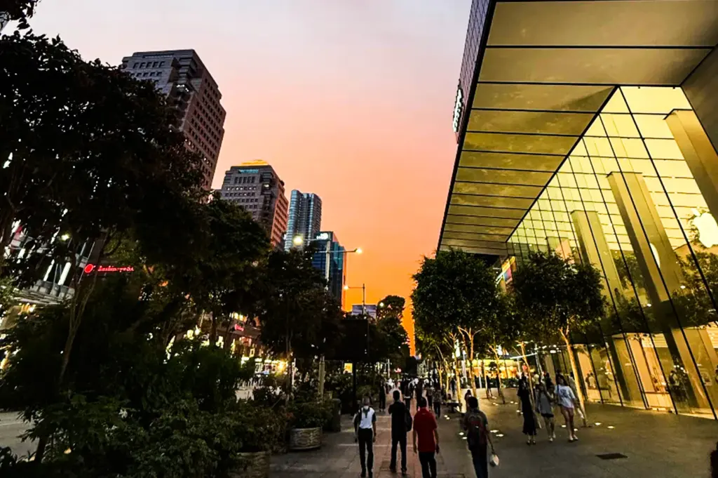 A wide street view of a bustling city sidewalk lined with lush green trees at sunset, featuring the glowing, multi-story glass facade of an Apple Store on the right and tall skyscrapers silhouette against a vibrant orange and pink sky in the background.