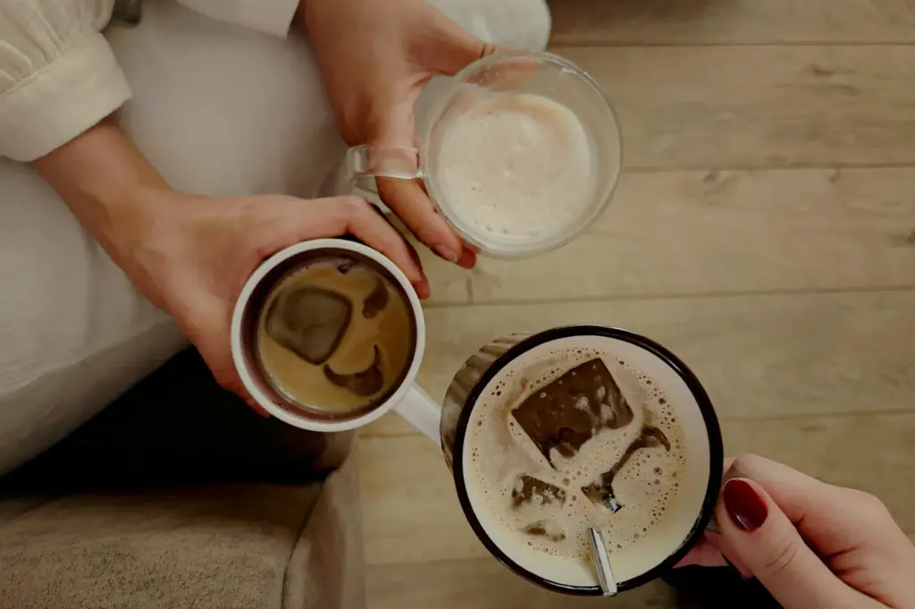 A top-down view of three people bringing their coffee mugs together for a toast over a light wooden floor. The drinks include two iced coffees with large square ice cubes and one foamy hot latte in a clear glass mug.