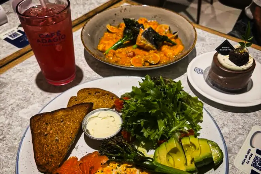 An overhead shot of a café meal laid out on a light marble table. In the foreground, a large white plate holds thick slices of toasted brown bread, a small cup of butter, scrambled eggs, smoked salmon, sliced avocado, grilled broccolini, and a fresh green salad. Behind it sits a shallow bowl filled with a creamy, orange-sauced pasta dish topped with seafood and broccolini. To the left is a tall glass of pink iced drink labeled "CAFÉ ADAGIO," and to the right is a small chocolate mousse cake on a white saucer.