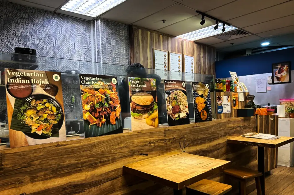 A cozy vegetarian restaurant interior with wooden tables and stools arranged along a long counter. Large menu display boards featuring dishes such as vegetarian rojak, char kway teow, vegan nasi lemak burger, and Hokkien mee line the counter, while warm lighting and wood-paneled decor create a casual and inviting dining atmosphere.