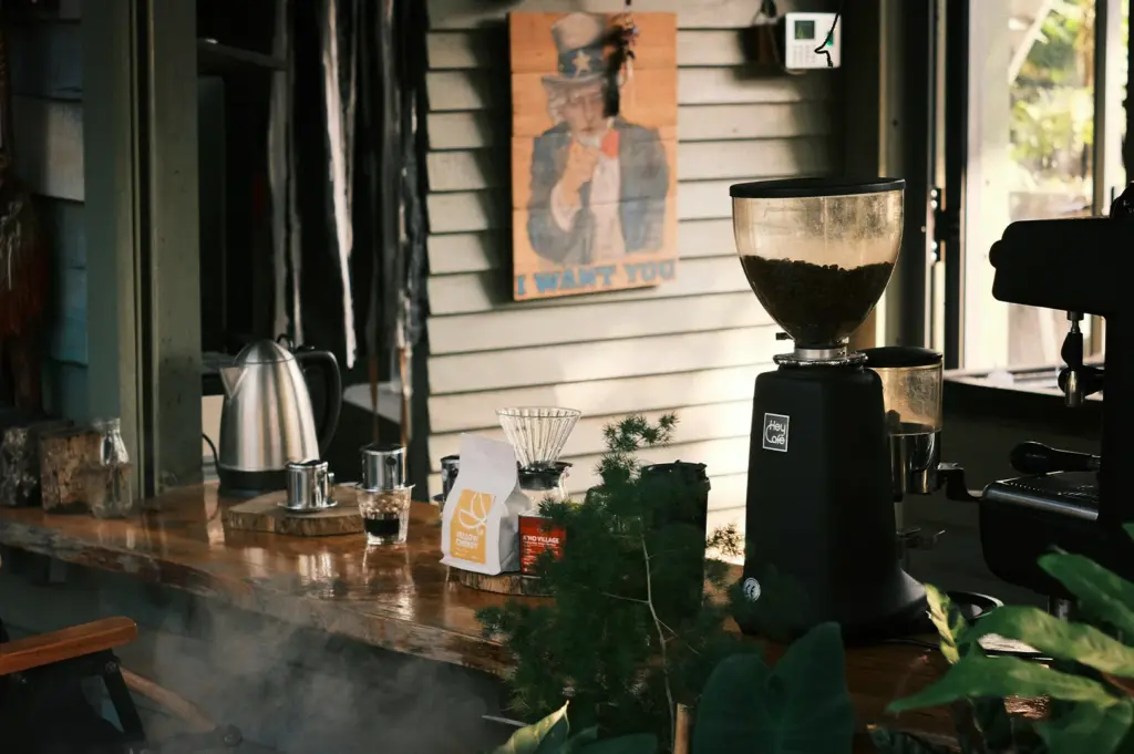 A wooden coffee bar setup featuring a black commercial coffee grinder filled with beans, pour-over equipment, a bag of "Yellow Cherry" coffee, and an electric kettle. A vintage "I Want You" Uncle Sam poster hangs on the light green paneled wall behind the counter.