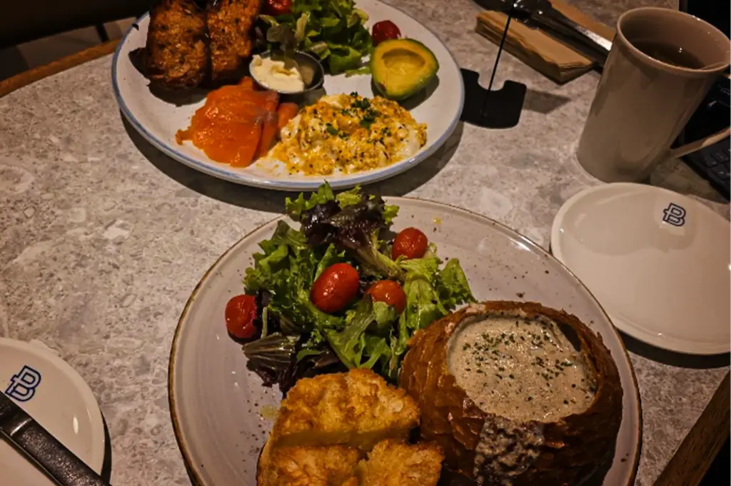 An eye-level shot of two brunch dishes served on a light marble table. In the foreground, a round grey plate features a bread bowl filled with creamy mushroom soup, accompanied by toasted bread pieces and a green salad with cherry tomatoes. In the background, another plate holds a breakfast spread with scrambled eggs, smoked salmon, avocado, toast, and salad. A white coffee mug and small saucers rest to the right.