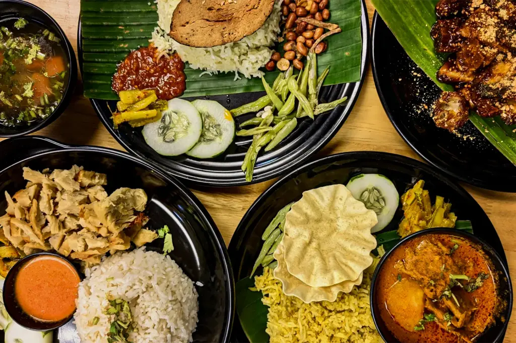 An overhead shot of a vibrant spread of Southeast Asian dishes, including Nasi Lemak with sambal, a rich curry set with yellow rice and papadum, shredded mock meat rice, and a side of rojak, all served on black plates lined with banana leaves.