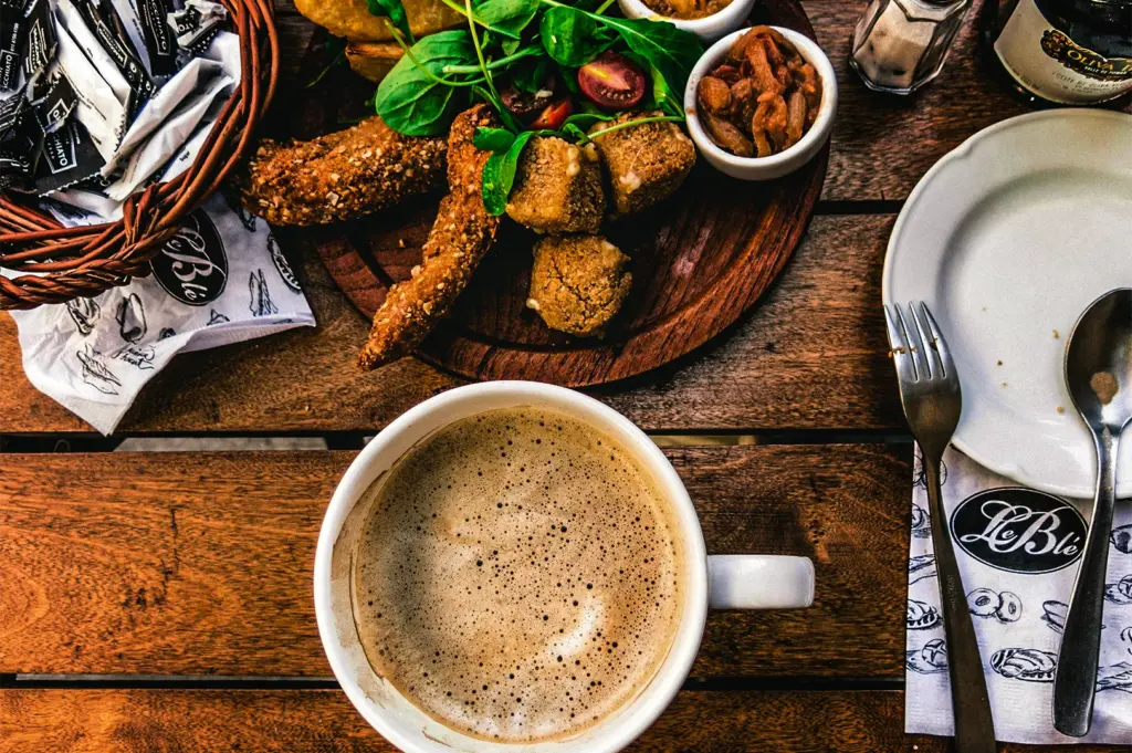 A top-down view of a rustic wooden table featuring a large, frothy cappuccino in a white mug, a wooden platter with fried finger foods, a salad, small condiment bowls, an empty white plate with a fork and spoon, and a small bread basket.