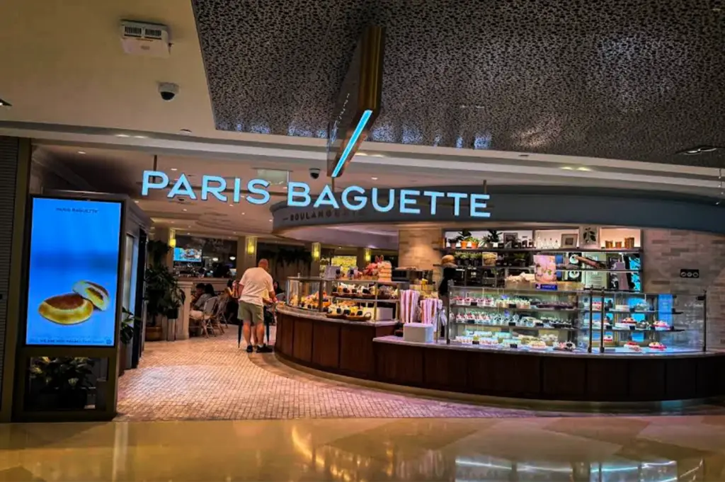 A wide shot of a curved "PARIS BAGUETTE" storefront situated inside a modern mall or commercial space with glossy floors. The glowing blue illuminated sign sits above an expansive, curved wooden counter equipped with large glass display cases showcasing various pastries and cakes. To the left, a tall digital poster displays an image of a pastry, while a male customer in shorts and a white t-shirt stands near the ordering area.