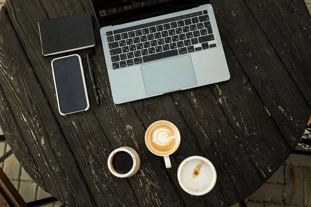 An overhead view of a dark wooden table arranged with an open laptop, a smartphone, a small black notebook with a pen, and three different coffee drinks including a latte with rosette latte art, a black coffee, and a frothy cappuccino.