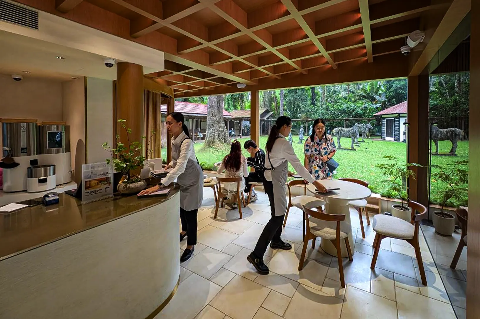 An eye-level, wide shot captures the elegant and serene dining room of a modern patisserie, representing the file "1-Ami Patisserie Orchard Elegant French-Japanese Pastries.webp". The interior features a warm, geometric wooden grid ceiling, polished stone floor tiles, and a curved service counter on the left where a staff member in a white shirt and grey apron stands ready. In the center of the room, another staff member prepares a white pedestal table surrounded by wooden cushioned chairs, while patrons chat at a table nearby. The entire space is brightened by expansive floor-to-ceiling windows that reveal a lush, sunlit green courtyard outside, complete with manicured grass, mature trees, and decorative horse sculptures, creating a peaceful, upscale ambiance.