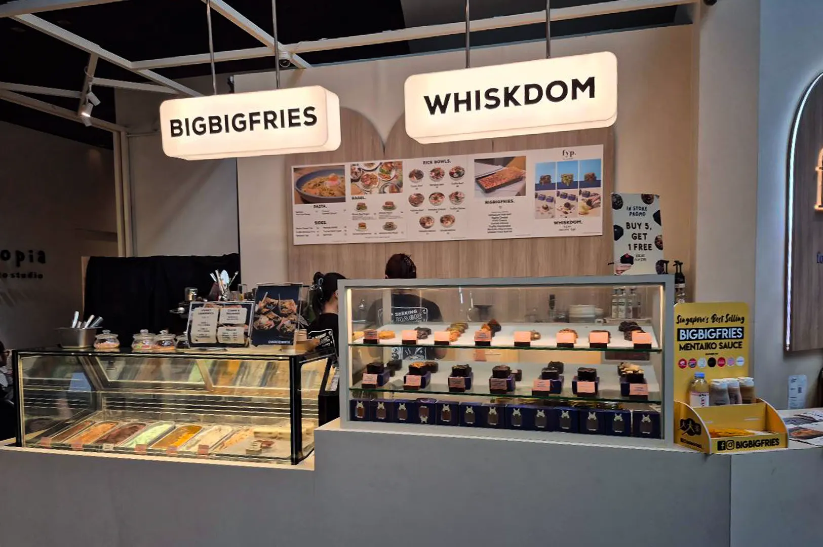 An eye-level shot captures a brightly lit indoor food kiosk hosting dual culinary concepts, representing the file "1-FYP Cafe Orchard Your Ultimate Destination.webp". Two large, glowing rectangular signs reading "BIGBIGFRIES" and "WHISKDOM" hang overhead above a light wood menu board that displays various pasta, bagel, and rice bowl options. The modern white service counter features a glass gelato display case on the left filled with colorful flavors, alongside a multi-tiered glass display case on the right showcasing neatly arranged rows of brownies and baked goods. Staff members in dark shirts work in the background, surrounded by promotional marketing materials, including a prominent yellow stand highlighting a "Mentaiko Sauce" blend, creating a clean and appealing casual dining setup.