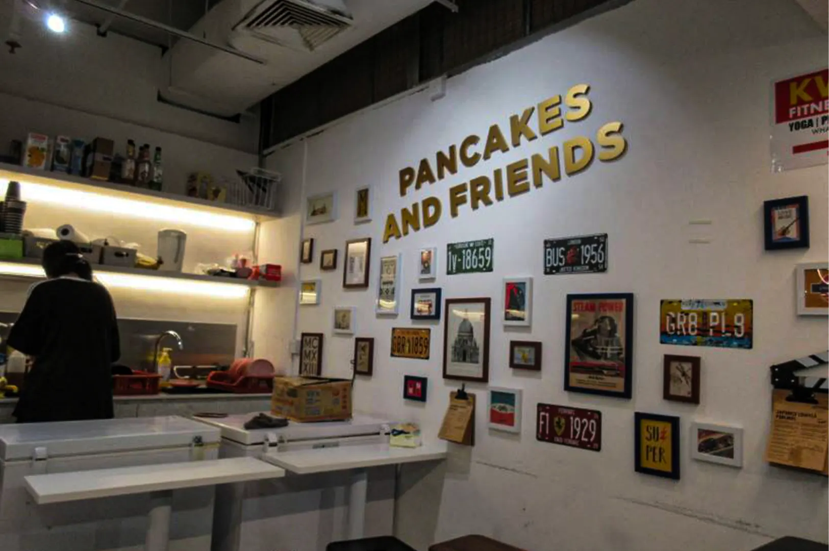 An eye-level shot captures the quirky, retro-inspired interior of a cafe kitchen and order area, representing the file "1-Pancakes And Friends Fluffy Pancakes.webp". On the right, a large white wall features the cafe's name, "PANCAKES AND FRIENDS," rendered in bold, 3D gold lettering, surrounded by a gallery arrangement of vintage license plates, framed architectural sketches, pop art prints, and small clipboards. To the left, a staff member with their back turned works behind a service station equipped with white counter tables and deep freezers, while dimly lit open shelving in the background holds kitchen ingredients, cups, and appliances under a warm light strip.