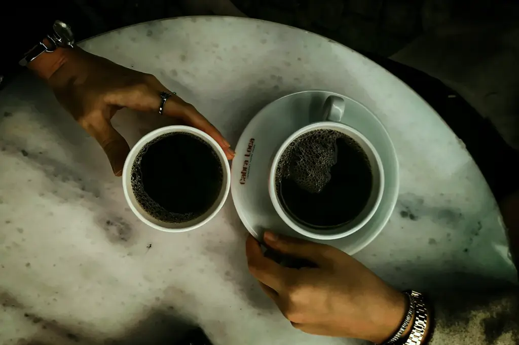 An overhead, top-down view of two people holding black coffee cups on a round, white marble table. On the left, a hand wearing a ring and a watch holds a simple white paper cup filled with coffee. On the right, another hand wearing a gold link bracelet holds a white ceramic cup and saucer, which has the red text "Cabra Loca" printed on the rim. Both cups contain dark coffee with slight bubbling on the surface.