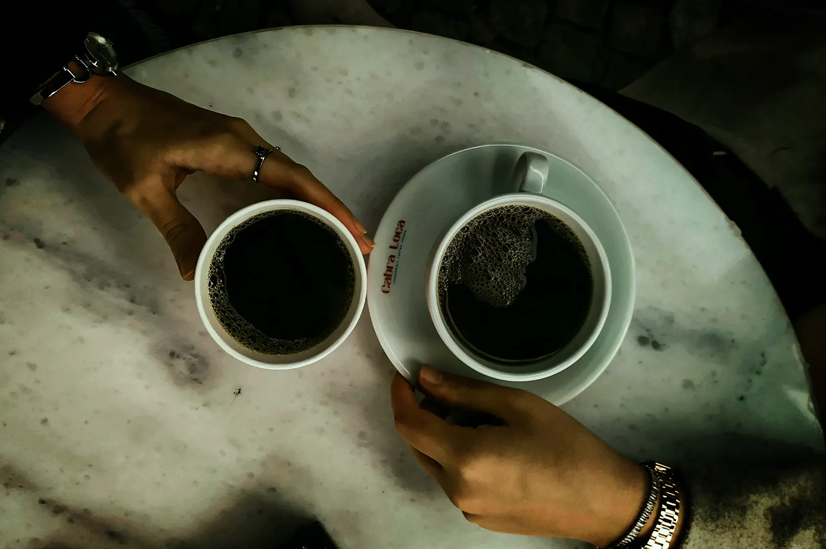 An overhead, top-down view of two people holding black coffee cups on a round, white marble table. On the left, a hand wearing a ring and a watch holds a simple white paper cup filled with coffee. On the right, another hand wearing a gold link bracelet holds a white ceramic cup and saucer, which has the red text "Cabra Loca" printed on the rim. Both cups contain dark coffee with slight bubbling on the surface.