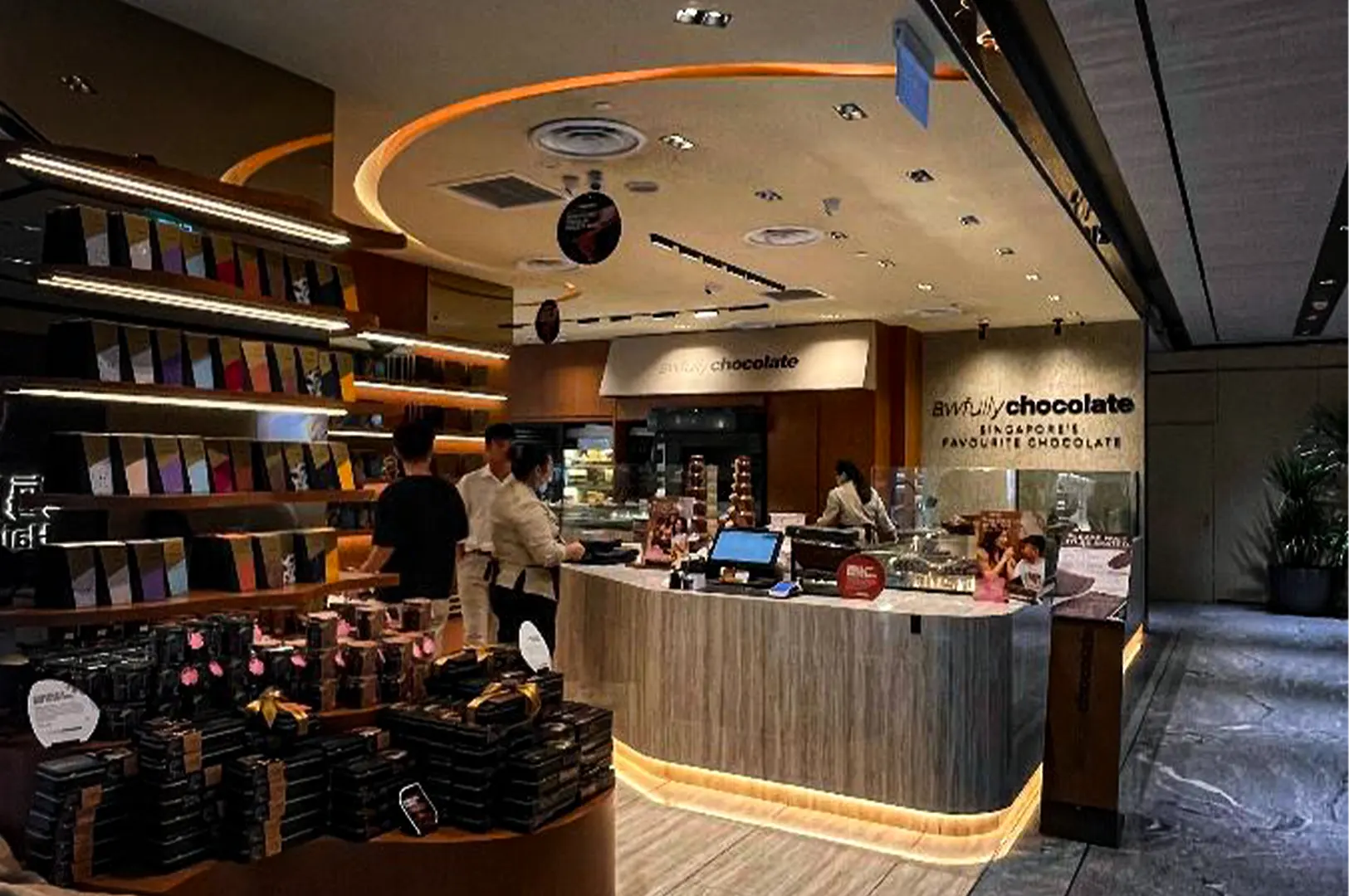 An eye-level shot captures the sleek, warm interior of a premium boutique chocolate store, representing the file "1-The Awfully Chocolate Experience.webp". Located in an indoor retail space with stone floors, the storefront features a prominent curved service counter wrapped in a light wood grain finish, highlighted by soft LED strip lighting along its base, where staff interact with customers near a digital point-of-sale monitor. To the left, a large tiered display island and illuminated wall shelves are meticulously stocked with rows of packaged chocolate gifts and boxes. The back wall features the bold signage "awfully chocolate SINGAPORE'S FAVOURITE CHOCOLATE," under a modern, circular-recessed ceiling illuminated by warm spotlights.
