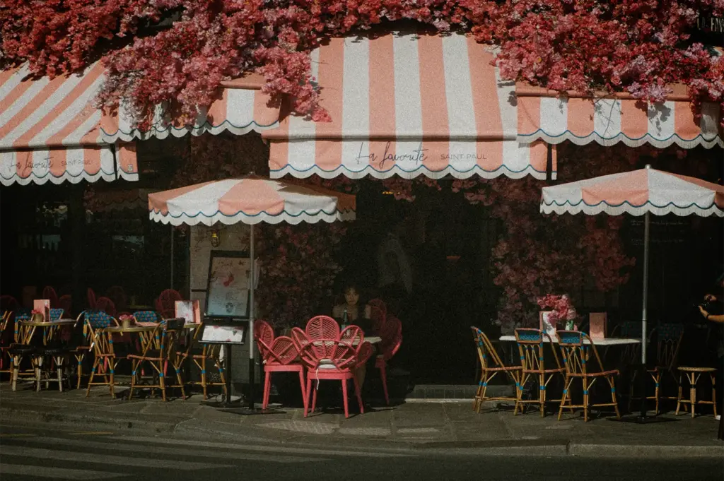 A charming, vintage-styled street view captures the storefront of a Parisian café named "La Favorite Saint-Paul," heavily adorned with dense clusters of pink flowers overhead and around the dark entrance. The café features distinctive pink-and-white striped awnings and matching patio umbrellas with scalloped edges. Below, an empty outdoor seating area is arranged along the stone sidewalk, complete with a mix of blue-accented wicker chairs and bright pink, fan-backed chairs gathered around small tables.