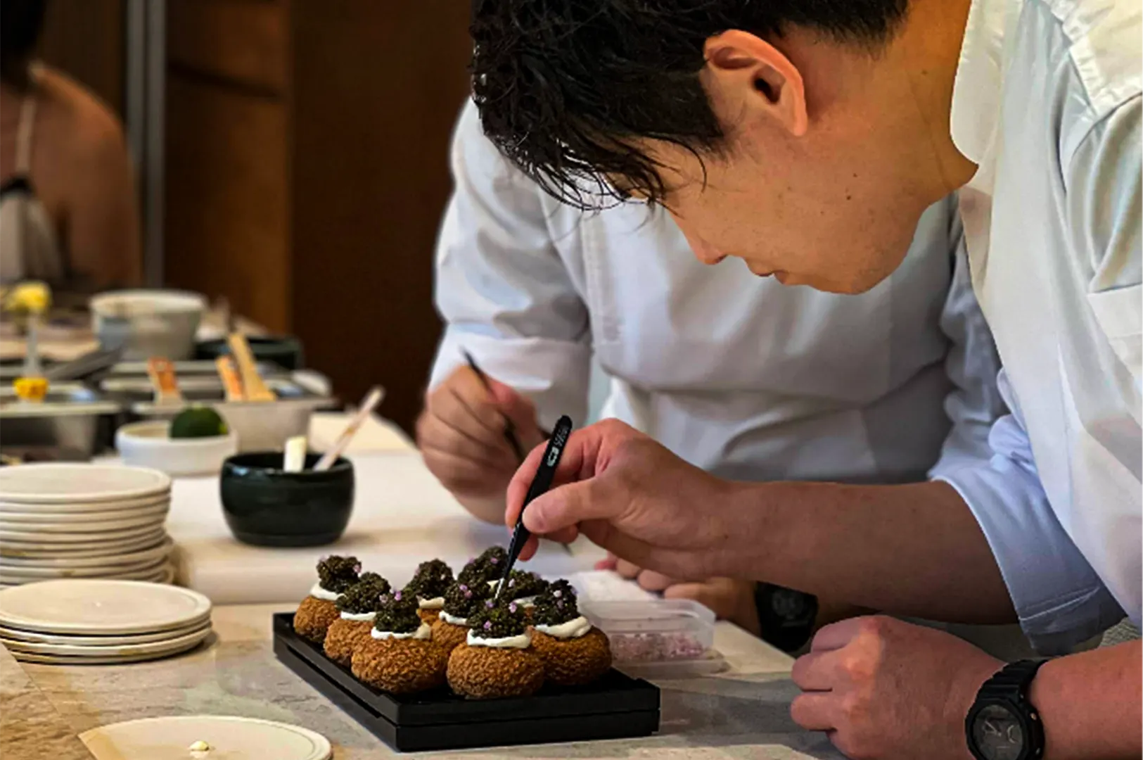 A close-up, profile shot captures the intense focus and precision of culinary professionals at work, representing the file "2-Ami Patisserie Orchard Elegant French-Japanese Pastries.webp". In the foreground, a pastry chef in a white uniform leans over a black tray, using a pair of fine black culinary tweezers to meticulously place a tiny purple flower garnish onto a row of delicate, round choux pastries topped with white cream and dark mounds of caviar or gelée. A second chef works closely alongside him in the softly blurred background, surrounded by a clean kitchen counter setup with small prep bowls, stacked white plates, and ingredient containers that emphasize the elegant, high-end pastry assembly process.