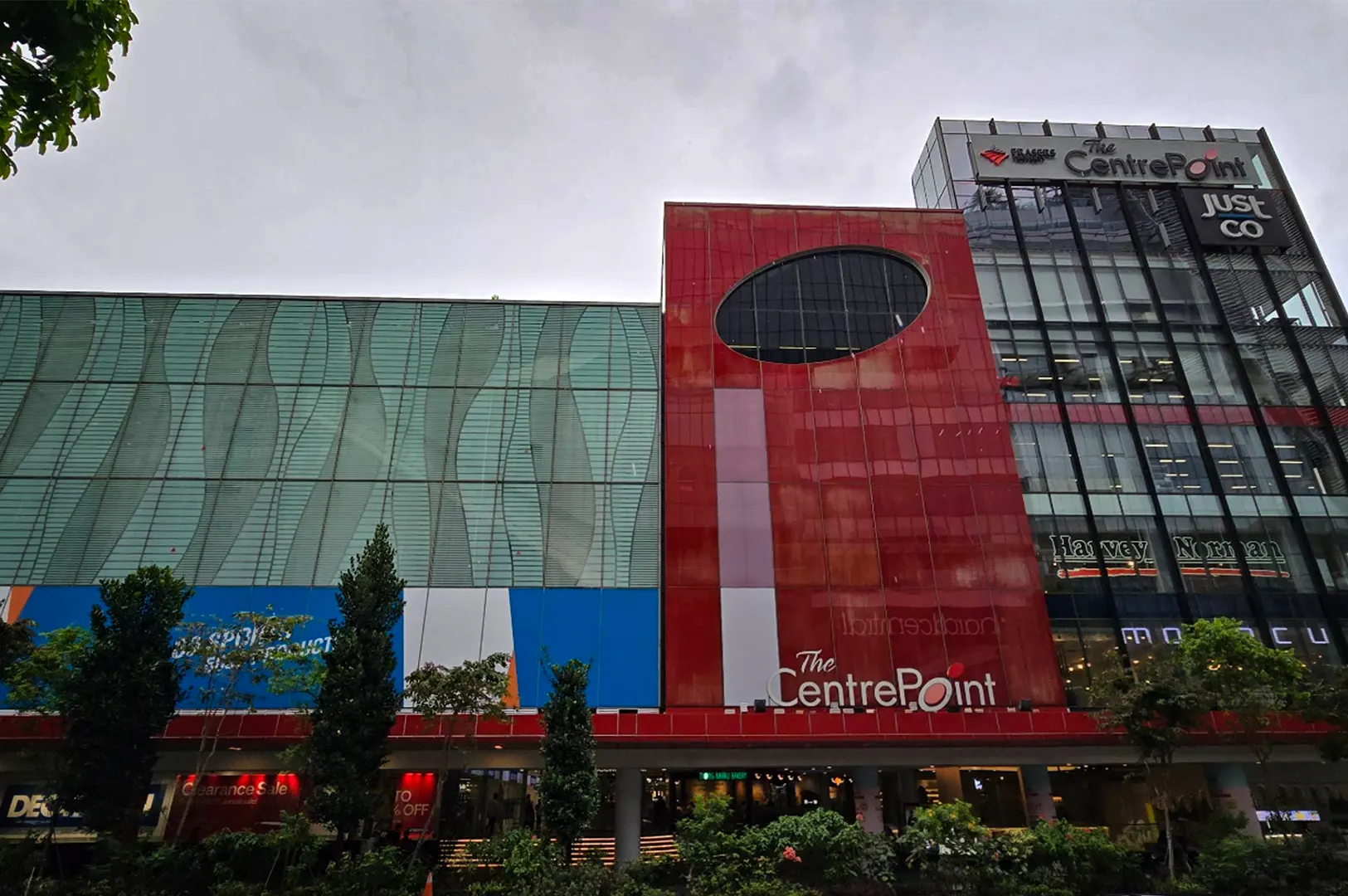 A low-angle, wide shot captures the modern, multi-textured exterior facade of a commercial shopping mall, representing the file "2-FYP Cafe Orchard Your Ultimate Destination.webp". The building's architecture is split into three distinct vertical sections under an overcast sky: a left wing covered in a light-green glass facade with wave-like vertical patterns, a prominent center block clad in smooth red panels featuring a large oval cutout window and "The CentrePoint" logo near the base, and a right wing consisting of a sleek glass-walled structure displaying tenant logos like "JustCo" and "Harvey Norman." Lush green trees and trimmed shrubbery line the ground-level walkway in the foreground, framing the vibrant urban retail landscape.