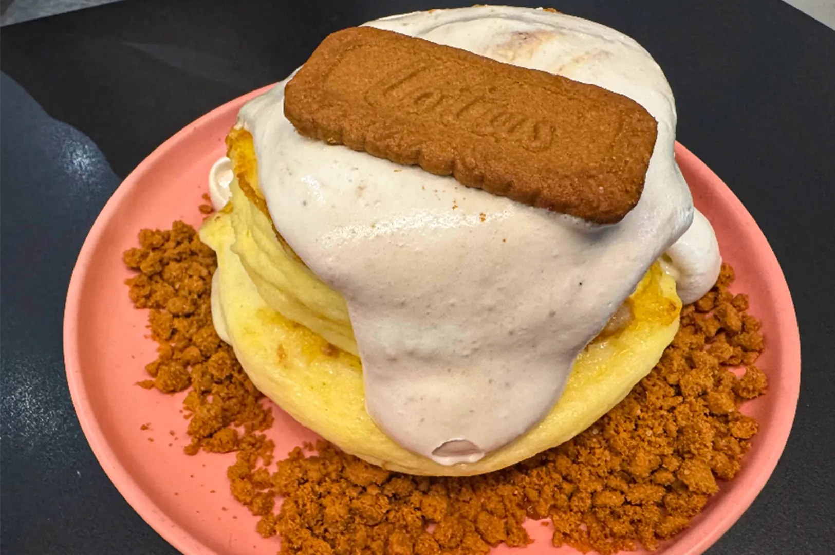 A high-angle, close-up shot features a decadent stack of thick, fluffy Japanese-style soufflé pancakes served on a matte pink plate. The pale golden pancakes are draped in a generous layer of thick, off-white cream that cascades down the sides, topped with a whole rectangular Biscoff speculoos cookie. Surrounding the base of the stack is a plentiful ring of crushed cinnamon-colored cookie crumbles, providing a textured contrast to the soft, airy pancakes against a dark, minimalist background.