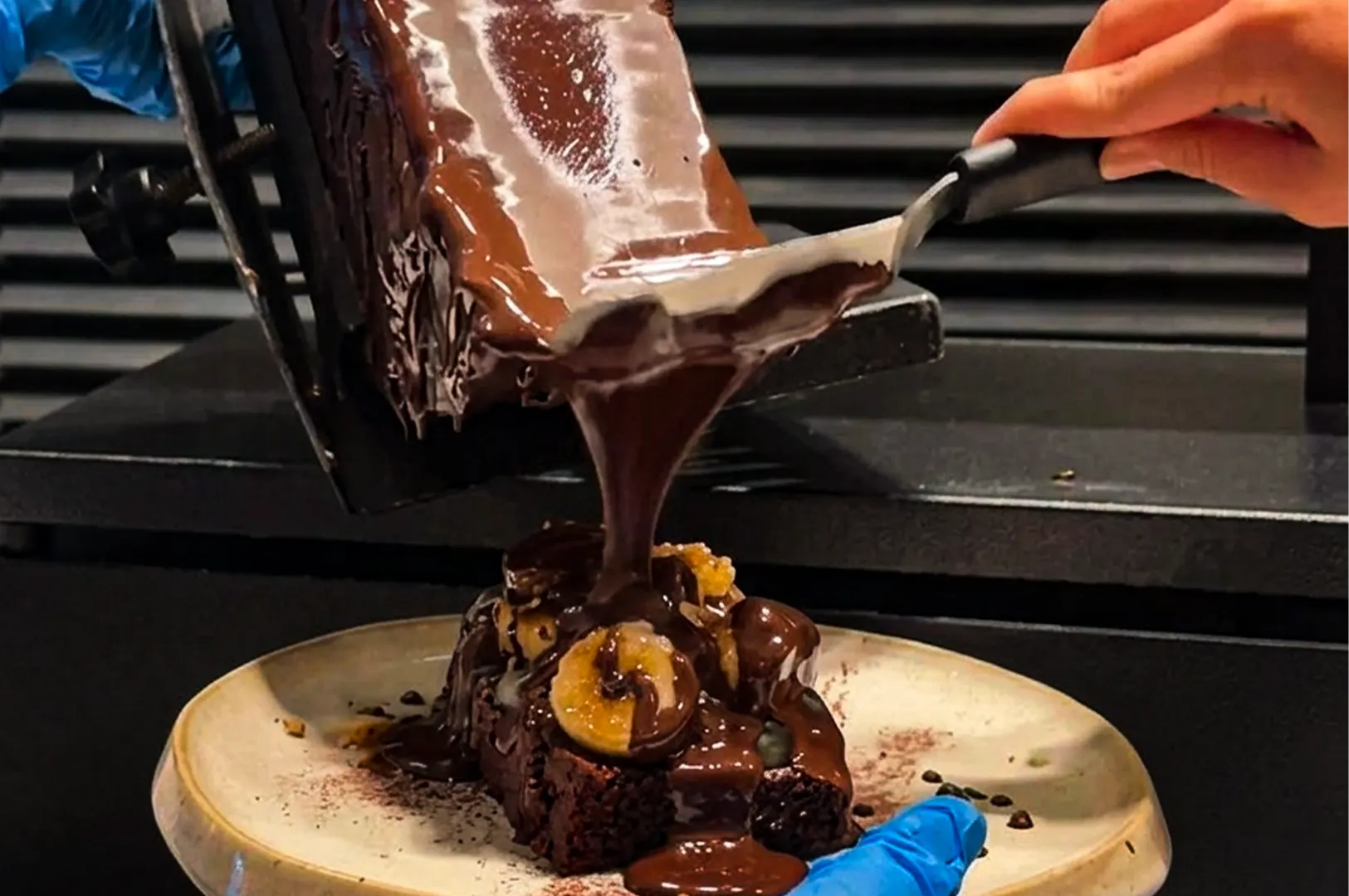 A close-up, action-oriented shot captures a decadent dessert being prepared by two individuals wearing blue gloves against a dark, slatted background. On the bottom of the frame, a person holds a speckled cream-colored plate featuring a thick, square dark chocolate brownie topped with sliced bananas. Simultaneously, another person uses a metal offset spatula to scrape a waterfall of glossy, melted dark chocolate from a vertical slab onto the brownie, creating a rich and indulgent coating. The warm lighting emphasizes the smooth texture of the flowing chocolate as it pools over the fruit and the dense cake base.