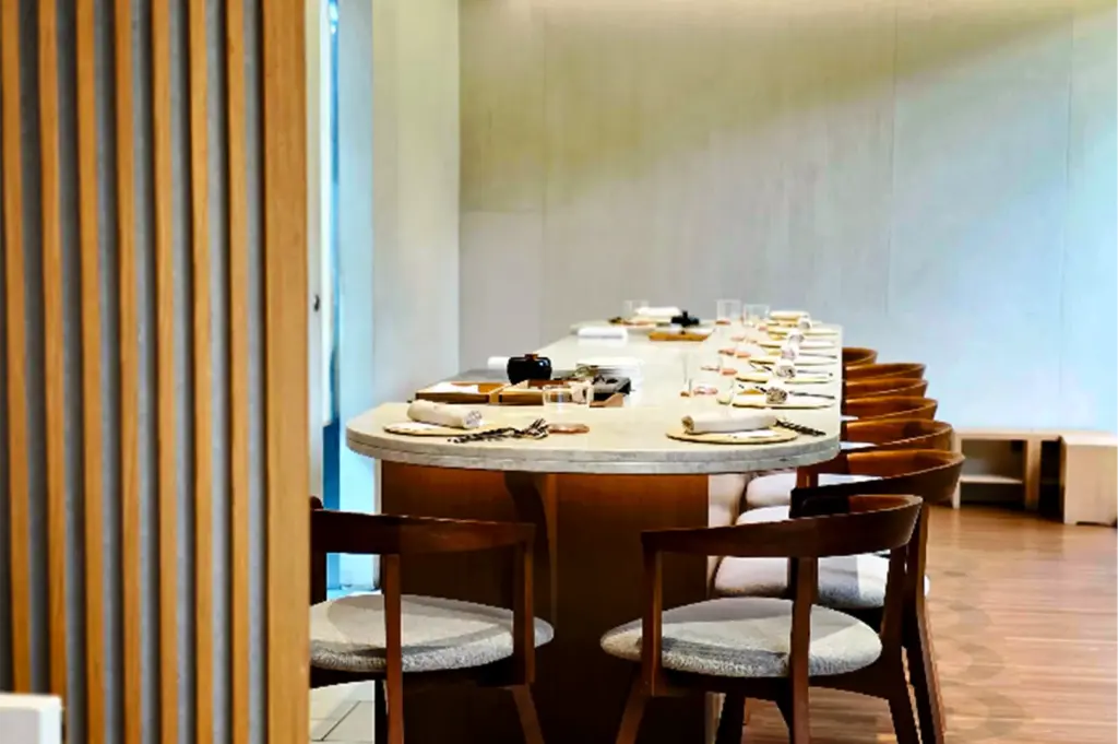 An eye-level, medium shot captures a minimalist and upscale chef's counter dining room, representing the file "3-Ami Patisserie Orchard Elegant French-Japanese Pastries.webp". A long, curved stone counter with a warm wooden base stretches diagonally across the space, fully set with rolled white napkins, water glasses, and small dishes for multiple guests. Elegant wooden chairs with cushioned light gray seats are neatly lined up along the counter, awaiting patrons. The serene ambiance is enhanced by a soft cream-colored back wall, polished wood flooring, and a vertically slatted wooden screen partially framing the left foreground, creating an intimate, high-end dining atmosphere.