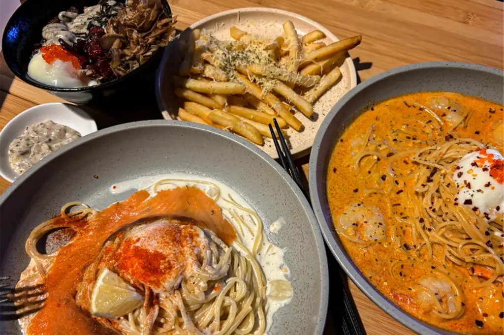 A high-angle shot captures an appetizing spread of fusion dishes arranged on a light wooden table, representing the file "3-FYP Cafe Orchard Your Ultimate Destination.webp". In the foreground, two large grey ceramic bowls feature pasta dishes: the one on the left contains noodles tossed in a creamy white sauce topped with smoked salmon, a soft-boiled egg dusted with red spice, and a lemon wedge, while the one on the right holds spaghetti in a rich, orange-tinted curry or laksa broth studded with shrimp and topped with a poached egg and chili flakes. Nestled between them in the background is a plate of golden French fries sprinkled with grated cheese and herbs alongside a dark bowl filled with a rice dish garnished with bonito flakes and fish roe, creating a rich and savory feast presentation.