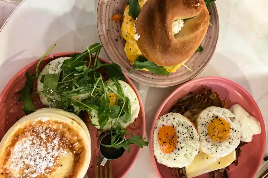 A overhead, flat-lay shot displays a hearty brunch spread featuring a variety of sweet and savory dishes arranged on a white table, representing the file "3-Pancakes And Friends Fluffy Pancakes.webp". On the bottom-left, a pink plate holds a stack of thick, fluffy pancakes dusted with powdered sugar, served alongside two fried eggs topped with a handful of fresh green arugula and a small metal pitcher of syrup. The top plate features a golden, glossy bagel sandwich overflowing with scrambled eggs, cheese, and greens, while the pink plate on the bottom-right presents a savory combination of crispy hash browns, melted cheese, two sunny-side-up eggs seasoned with black pepper, and a dollop of white cream, creating a colorful and inviting meal presentation.