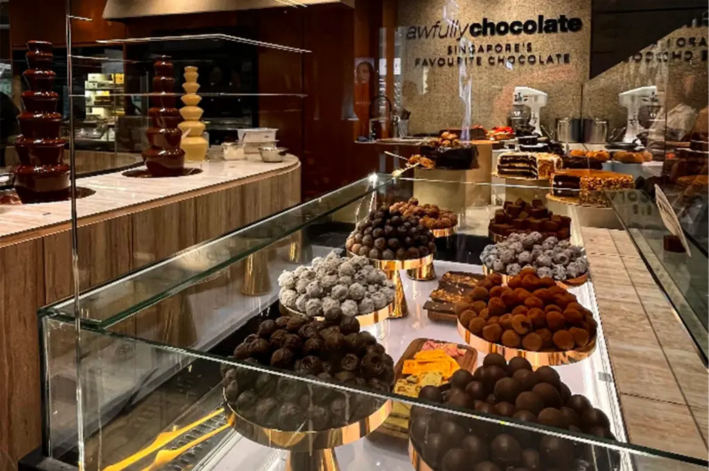 An eye-level shot captures a vibrant and luxurious chocolate display case inside a boutique dessert shop, representing the file "3-The Awfully Chocolate Experience.webp". In the foreground, a long, angled glass counter showcases an assortment of rich chocolate truffles, pralines, and confections arranged in generous, rounded mounds on elevated gold pedestals. In the mid-ground to the left, a curved wooden counter houses three operating multi-tiered chocolate fountains—two flowing with dark chocolate and one with white chocolate. The background reveals a selection of decadent gourmet cakes on stands, kitchen equipment like stand mixers, and the store's signature signage "awfully chocolate SINGAPORE'S FAVOURITE CHOCOLATE" printed on a textured beige wall, completing the indulgent, high-end culinary atmosphere.