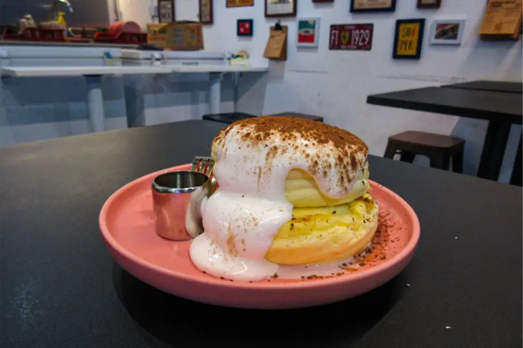 An eye-level close-up shot captures a decadent stack of thick, fluffy Japanese-style soufflé pancakes resting on a matte pink plate on a dark table, representing the file "4-Pancakes And Friends Fluffy Pancakes.webp". The two golden, airy pancakes are layered together and generously draped in a thick, velvety white cream that cascades down the side, heavily dusted on top with rich cocoa powder. Next to the pancakes sits a small, metallic pitcher of syrup with a fork resting behind it. In the softly blurred background, the cozy cafe interior is visible, featuring a white wall adorned with eclectic vintage license plates and framed art prints, empty dark dining tables, and white service counter preparation tables.