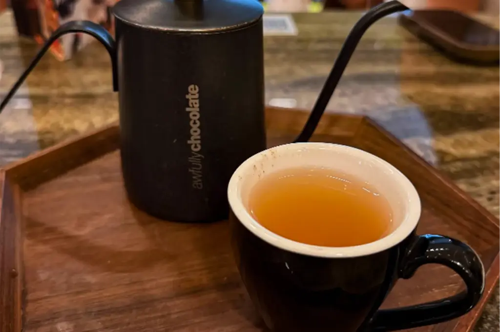 A close-up, slightly high-angle shot captures a warm beverage service arrangement resting on a hexagonal wooden tray, representing the file "4-The Awfully Chocolate Experience.webp". In the foreground, a glossy black ceramic cup with a white interior is filled with a translucent, amber-colored tea. Behind and slightly to the left of the cup sits a matte black, cylindrical mini gooseneck kettle with its slender, curved spout extending upward and to the right. The side of the kettle is subtly branded with the vertical white text "awfullychocolate". The entire tray rests on a polished, darkly marbled tabletop that creates a softly blurred, sophisticated background.