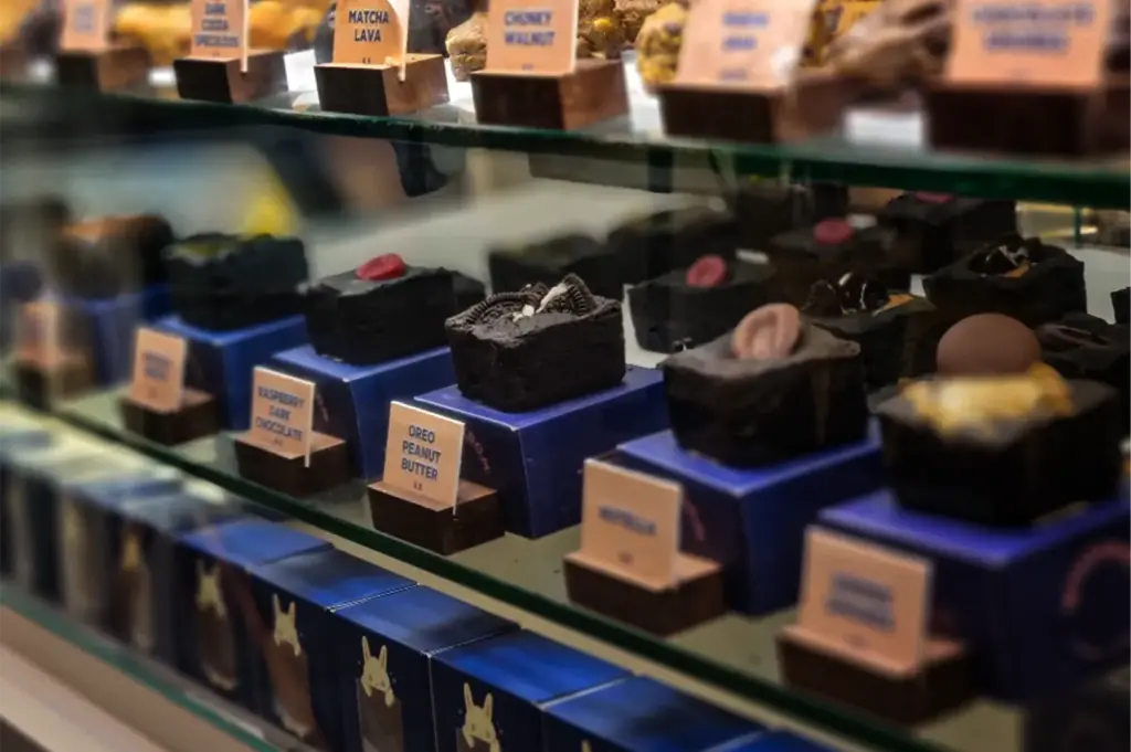A close-up, angled shot captures a multi-tiered glass bakery display case filled with gourmet baked treats, representing the file "5-FYP Cafe Orchard Your Ultimate Destination.webp". The middle glass shelf features an orderly row of dense, dark chocolate brownie blocks neatly positioned on top of individual dark blue boxes, each accompanied by small wooden menu labels such as "Raspberry Dark Chocolate" and "Oreo Peanut Butter." The prominent central brownie is topped with crushed chocolate sandwich cookies, while adjacent treats are garnished with red berries and chocolate drizzles. The top and bottom shelves are softly blurred, showing a peek of cookies like "Matcha Lava" above and blue retail packaging featuring a small rabbit character lined up below.