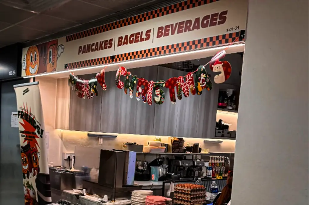 An eye-level shot captures the exterior storefront and open kitchen of a casual eatery, representing the file "5-Pancakes And Friends Fluffy Pancakes.webp". A prominent horizontal sign running across the top of the storefront reads "PANCAKES | BAGELS | BEVERAGES" in bold brown lettering, framed by an orange and black checkered pattern and vintage-style cartoon pancake illustrations. Strung directly underneath the sign is a colorful, festive "Merry Christmas" paper banner shaped like patterned holiday stockings. The open service counter reveals a busy kitchen area equipped with stacks of plates, commercial espresso machines, flavored syrup bottles, an industrial mixer, and multiple trays of brown eggs, while a tall vertical banner stands on the left featuring animated cartoon characters.