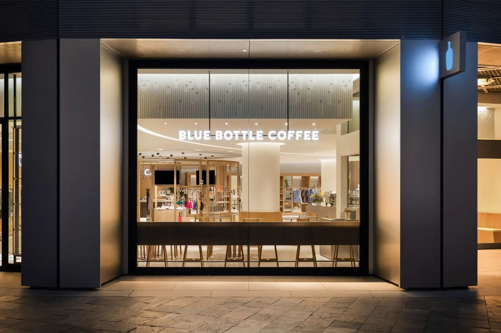 A straight-on exterior shot captures a modern Blue Bottle Coffee storefront with a massive glass window displaying a brightly lit, minimalist interior. The shop features clean lines, warm wooden seating counters, and illuminated white text signage above the seating area, flanked by thick architectural pillars.