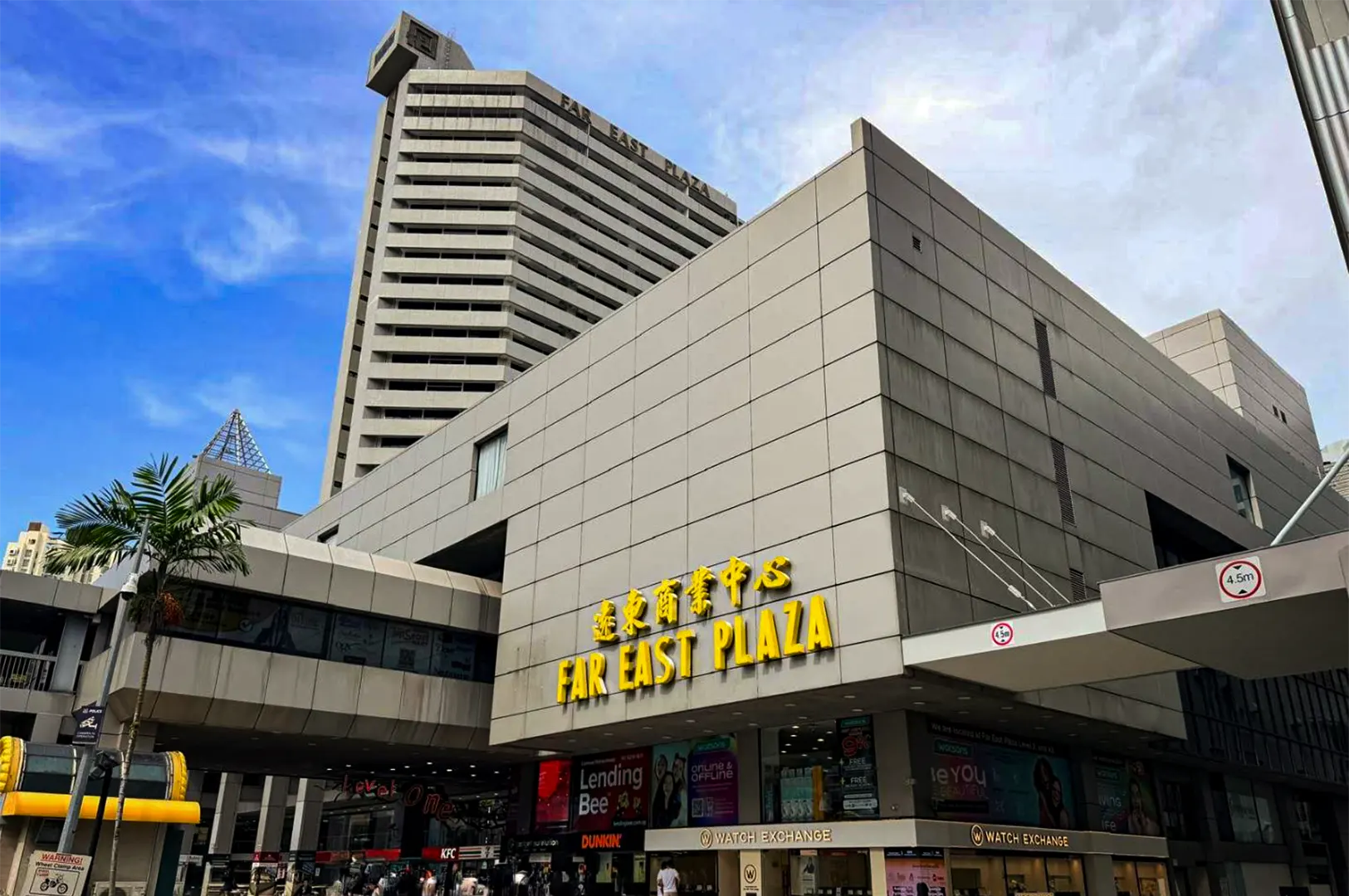 A low-angle, wide shot captures the exterior of Far East Plaza, a prominent multi-story commercial complex under a bright blue sky with scattered wispy clouds. The building's modernist architecture is characterized by a textured, grey-paneled square facade featuring large, bright yellow signage that reads "FAR EAST PLAZA" in both English and Chinese characters. Behind this main block, a taller, tiered concrete skyscraper with the words "FAR EAST PLAZA" visible near its upper rim towers into the sky. At the street level, a covered walkway and various storefront signs—including "Watch Exchange," "Lending Bee," and "Dunkin'"—line the base of the bustling shopping hub, while a single palm tree stands to the far left.