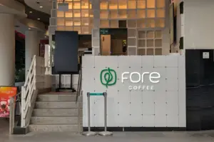 The modern storefront of Fore Coffee features white tiled walls with a prominent green leaf logo and white lettering. A small set of stairs leads up to the ordering and pick-up counter under glowing ceiling panel lights.