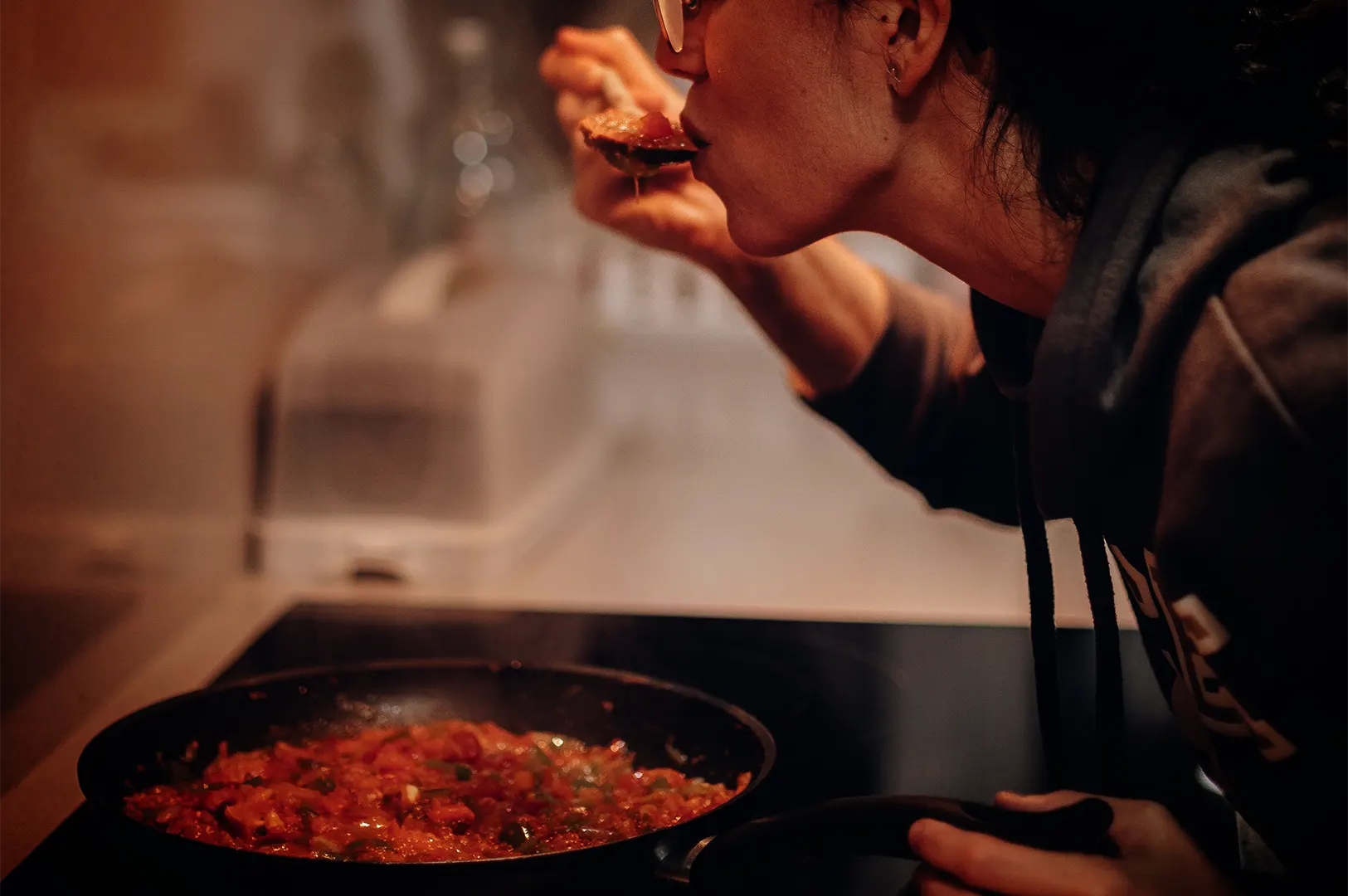 A close-up, profile shot captures a person with glasses and a dark hoodie tasting a simmering dish directly from a spoon in a dimly lit kitchen. Their face is angled downward as they gently blow on or sip a bite of food from a wooden spoon. In the lower-left foreground, a black frying pan rests on a smooth stovetop cooktop, filled with a colorful, chunky red sauce or stir-fry mixed with chopped green vegetables. The warm, moody overhead lighting casts soft shadows, creating an intimate atmosphere focused on the act of cooking.