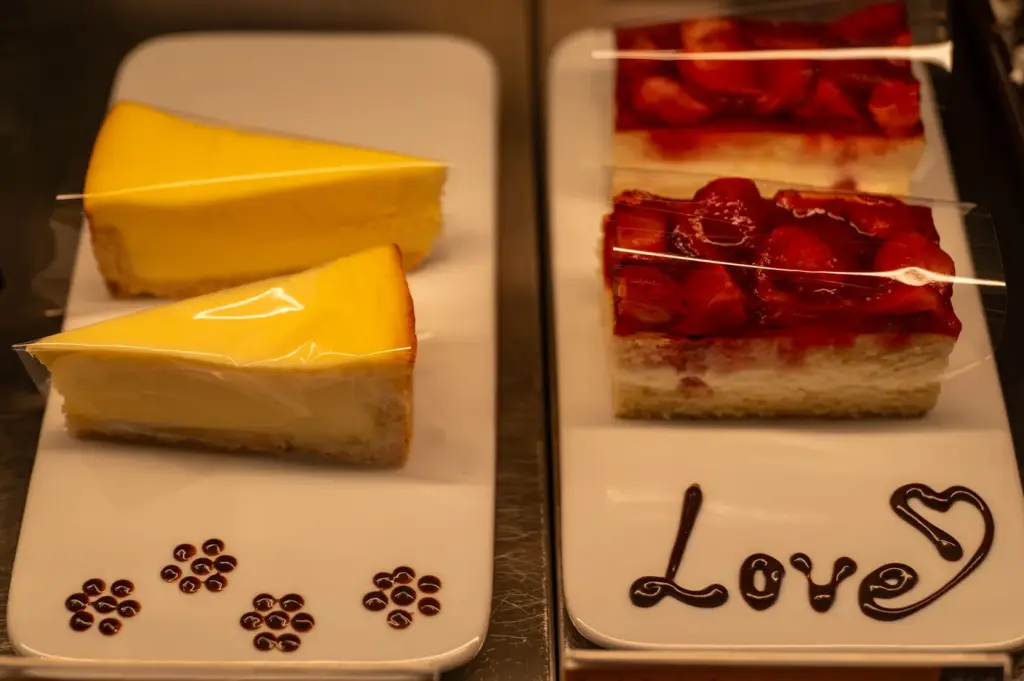 Slices of smooth lemon tart and layered strawberry cake are displayed on white rectangular trays inside a glass case. The trays are decorated with dark chocolate sauce, one featuring small paw-print dots and the other spelling out the word "Love" with a heart.
