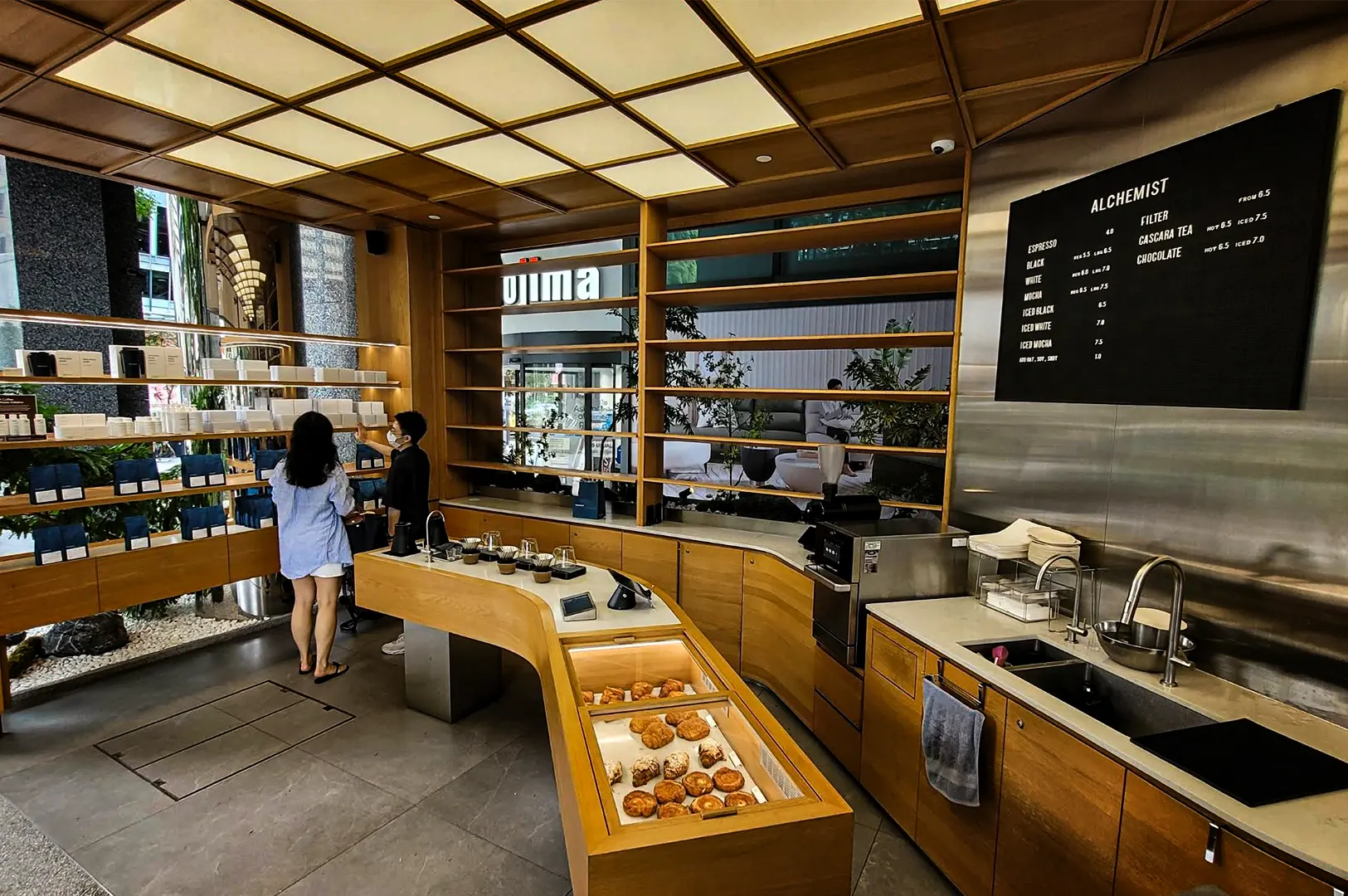 A high-angle, wide-set shot captures the minimalist and warm interior of Alchemist coffee shop. The space features light-toned wooden cabinetry and a distinctive gridded, illuminated ceiling canopy that bathes the cafe in bright, even light. A long, curved wooden service counter winds through the center, integrating a glass pastry display case filled with croissants, a pour-over coffee station, and a stainless steel sink setup on the right below a large black menu board. To the left, a customer in a blue shirt stands near retail shelves browsing coffee bags while a staff member in a black shirt stands nearby. In the background, floor-to-ceiling windows look out onto an open-air building courtyard with lush green plants.