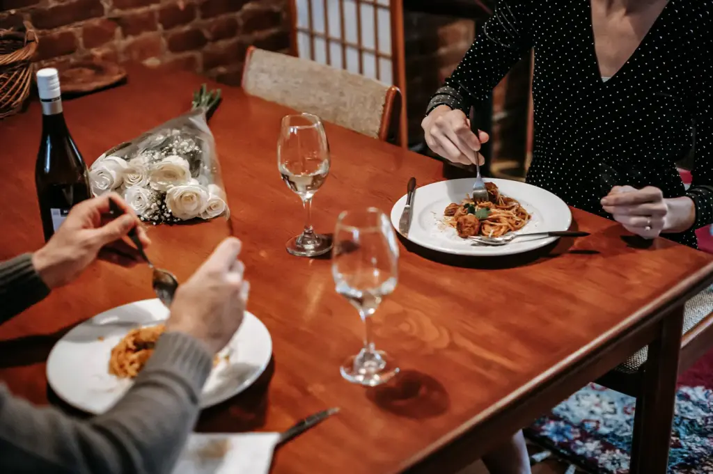 A high-angle shot captures a couple enjoying a romantic dinner date at a wooden table, both eating plates of spaghetti with meatballs. A bottle of wine, two wine glasses, and a bouquet of white roses sit on the table, setting an intimate mood in the warmly lit room.