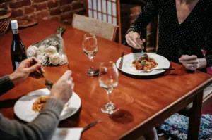A high-angle shot captures a couple enjoying a romantic dinner date at a wooden table, both eating plates of spaghetti with meatballs. A bottle of wine, two wine glasses, and a bouquet of white roses sit on the table, setting an intimate mood in the warmly lit room.