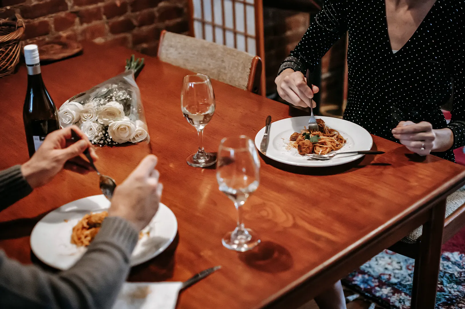 A high-angle shot captures a couple enjoying a romantic dinner date at a wooden table, both eating plates of spaghetti with meatballs. A bottle of wine, two wine glasses, and a bouquet of white roses sit on the table, setting an intimate mood in the warmly lit room.