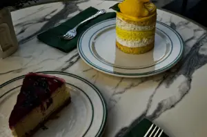 A high-angle shot captures two slices of dessert served on white plates with green borders, arranged on a marble tabletop. A slice of blueberry cheesecake sits in the foreground, while a layered lemon cake topped with a small, yellow lemon-shaped decoration rests in the background alongside silver forks and green napkins.