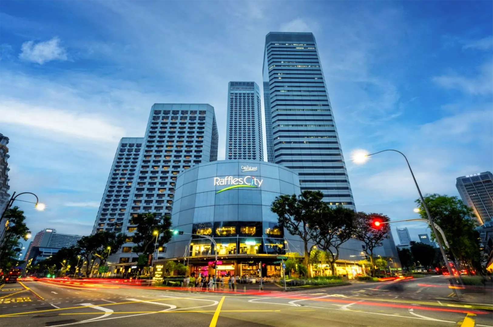 A low-angle, wide shot captures the modern, curved facade of the Raffles City shopping complex, flanked by towering skyscrapers under a blue twilight sky. In the foreground, long-exposure light trails from passing city traffic streak across the wide street intersection.