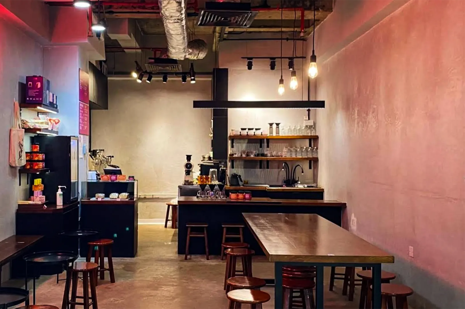 An eye-level, wide shot captures the moody, industrial interior of a specialty coffee shop with concrete walls bathed in a warm, pinkish-red ambient glow. In the foreground, a long wooden communal table with metal legs is surrounded by matching round wooden stools. Towards the back, a dark service counter is equipped with a pour-over station, a coffee grinder, and rows of glassware arranged neatly on open wooden shelves underneath hanging Edison bulb light fixtures. To the left, a smaller cash register counter features a glass pastry display case next to a retail corner showcasing coffee bags and merchandise, all set under an exposed ceiling with industrial pipes and track lighting.