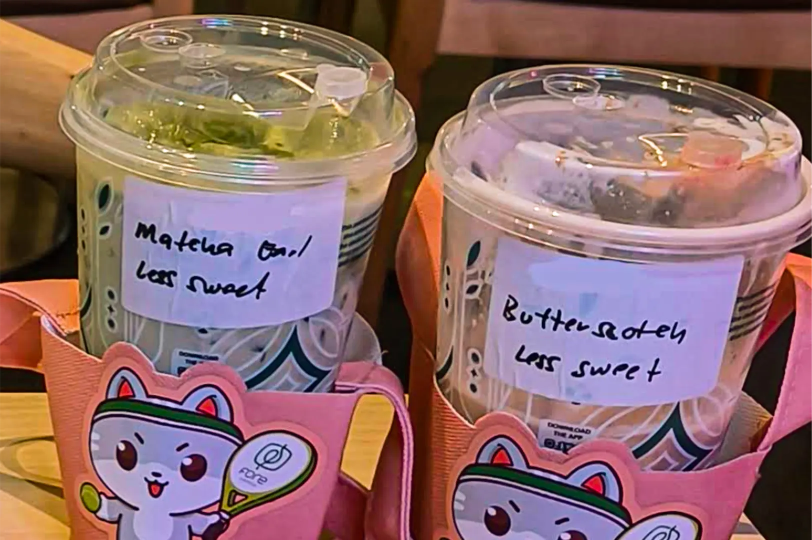 Two iced drinks in clear plastic cups are held in pink, cartoon-cat drink carriers with handles. Handwritten labels taped to the cups read "Matcha Oat less sweet" for the green tea beverage and "Butterscotch less sweet" for the iced coffee.