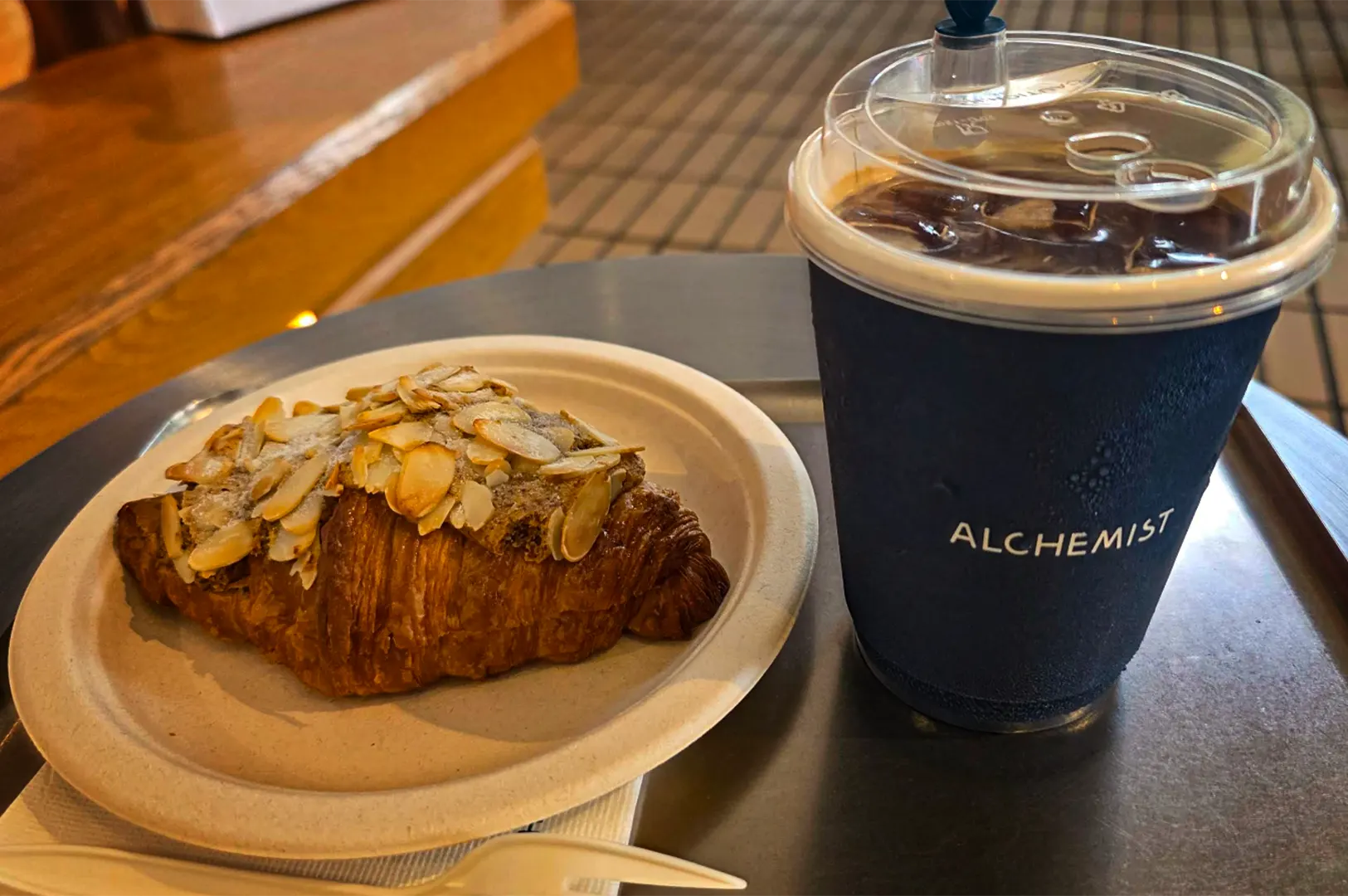 A close-up, slightly high-angle shot captures a morning coffee combination served on a grey tray resting on a dark table surface. To the left, a golden-brown almond croissant generously topped with sliced, toasted almonds and dusted with powdered sugar sits on a textured, off-white paper plate. To the right, a cold beverage, showing visible condensation, is served in a dark blue cup bearing the white text "ALCHEMIST," covered with a clear plastic domed lid that reveals ice cubes inside. In the foreground, a white plastic fork rests on a napkin next to the plate, while the background softly blurs into the warm wooden tones of an indoor dining space and tiled flooring.