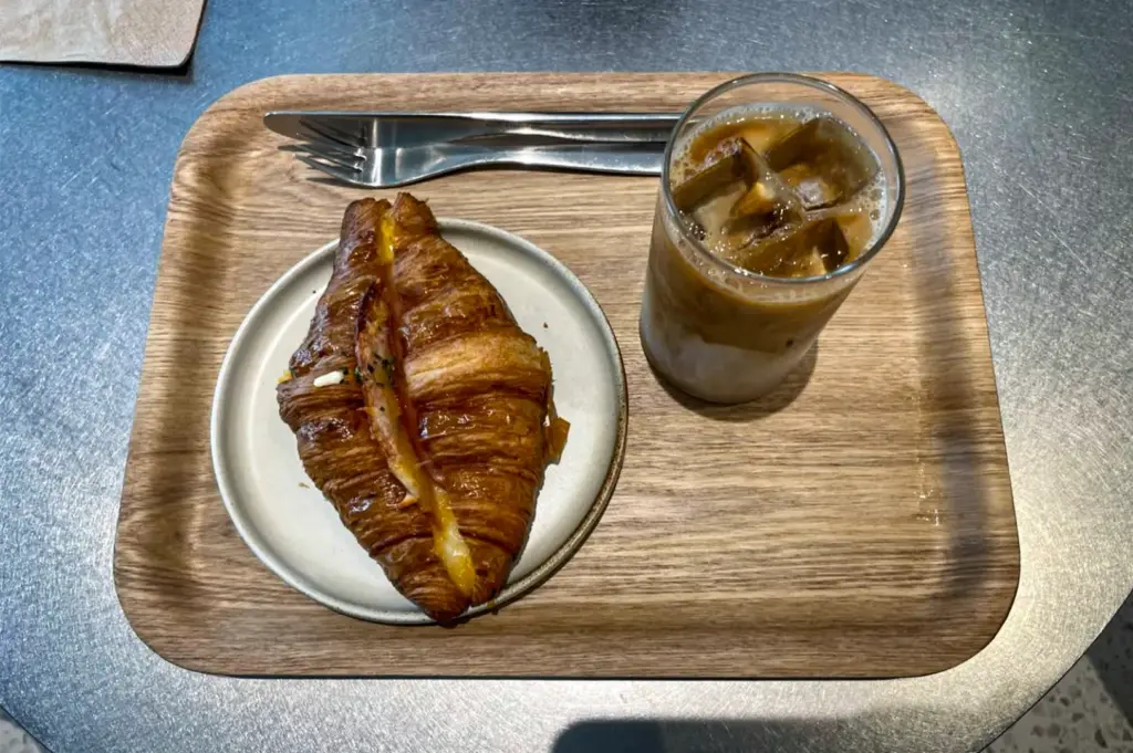 A high-angle shot captures a savory, golden-brown croissant split open with melted cheese on a small ceramic plate. It sits alongside a clear glass of iced latte showing distinct layers of milk and espresso on a rectangular wooden tray with a silver fork and knife.