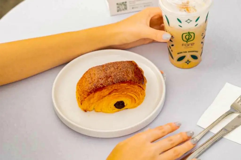 A golden-brown pain au chocolat sits on a round white ceramic plate next to an iced coffee drink in a branded Fore Coffee plastic cup. A person's hands reach toward the food and beverage on a clean white table set with silverware.