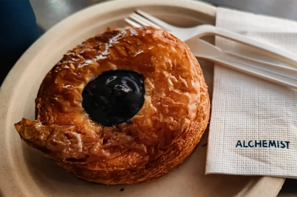 A close-up, high-angle shot captures a round, golden-brown Danish pastry served on a textured off-white paper plate. The pastry features crispy, flaky caramelized layers with a glossy, dark blue berry filling neatly centered in the middle. To the right, a white plastic fork and knife rest across a folded white paper napkin featuring the clean, blue printed text "ALCHEMIST," all presented under bright, natural light.