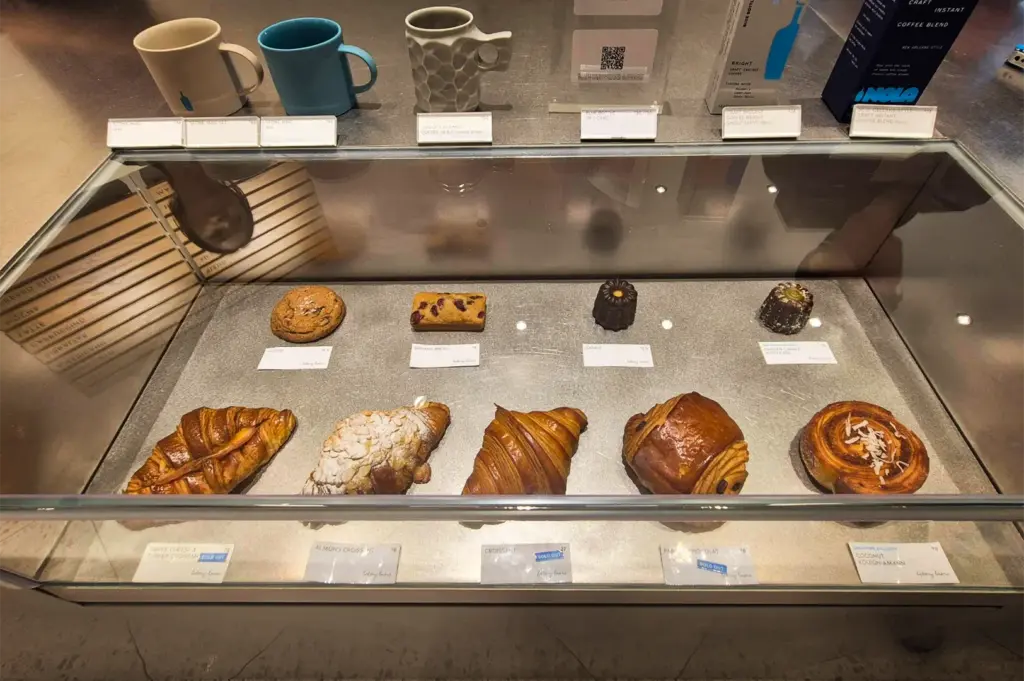 A high-angle shot displays a glass bakery case filled with pastries, including multiple golden-brown croissants, a cookie, a small loaf, and canelés arranged on a gray surface with neat white price labels. Above the glass display, a collection of merchandise including ceramic coffee mugs, a QR code sign, and boxed instant coffee blends are lined up on the counter.