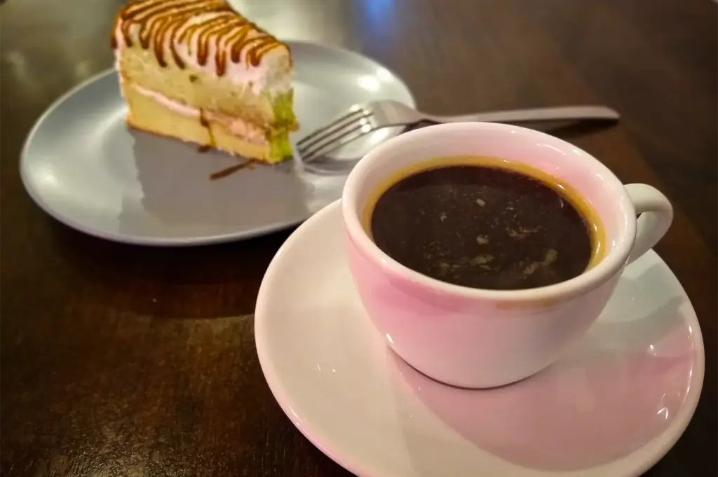 A close-up, slightly high-angle shot captures a comforting coffee and dessert pairing served on a dark wooden table. In the foreground, a white ceramic cup filled with rich, dark espresso displaying a thin layer of crema sits on a matching white saucer. Softly blurred in the background, a slice of layered sponge cake with cream frosting and a drizzle of caramel syrup is presented on a light grey plate, accompanied by a silver metal dessert fork resting alongside it.