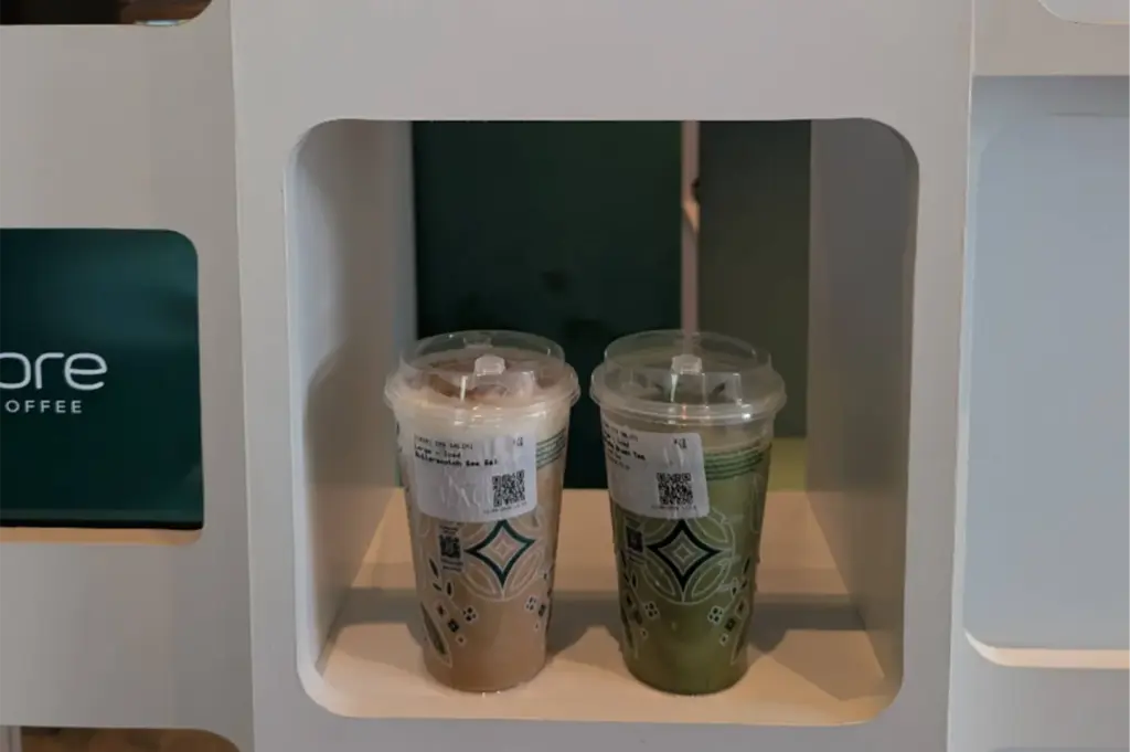 Two iced beverages in clear, patterned plastic cups sit inside a white rectangular pick-up shelf at a Fore Coffee store. One cup holds a layered iced butterscotch coffee with foam topping, while the other contains a green matcha tea, both featuring printed order labels with QR codes.