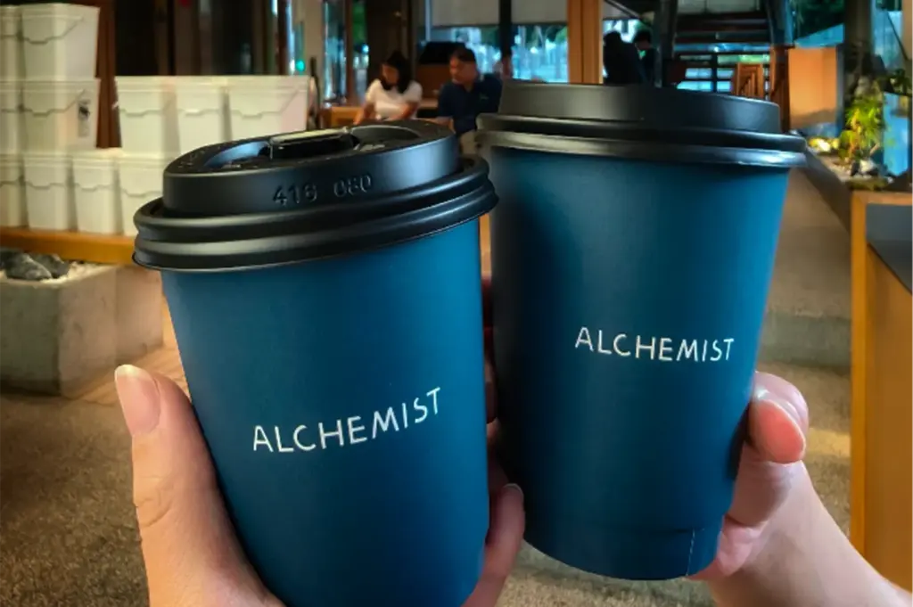 A close-up, eye-level shot captures two hands holding up a pair of takeaway coffee cups side by side in a toast-like gesture. The matching textured paper cups are a deep teal color, each featuring the white printed name "ALCHEMIST" across the front and capped with a black plastic travel lid. In the dimly lit, softly blurred background, the interior of the coffee shop unfolds with warm wood tones, a stone planter on the left, stacks of white buckets or containers, and a couple of patrons seated at tables under soft evening light.